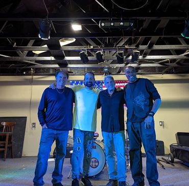 Four men smiling on a stage with musical instruments behind them.