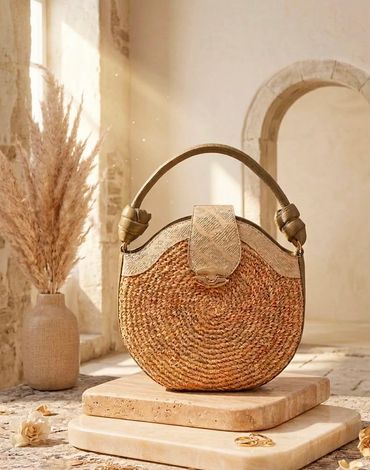 Round woven straw handbag with leather handle in a sunlit rustic setting.
