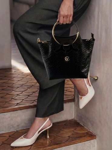 Elegant black handbag with gold handles paired with white high-heeled shoes and dark pants.