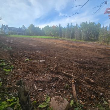 A cleared dirt field with a house and trees in the background under a partly cloudy sky.