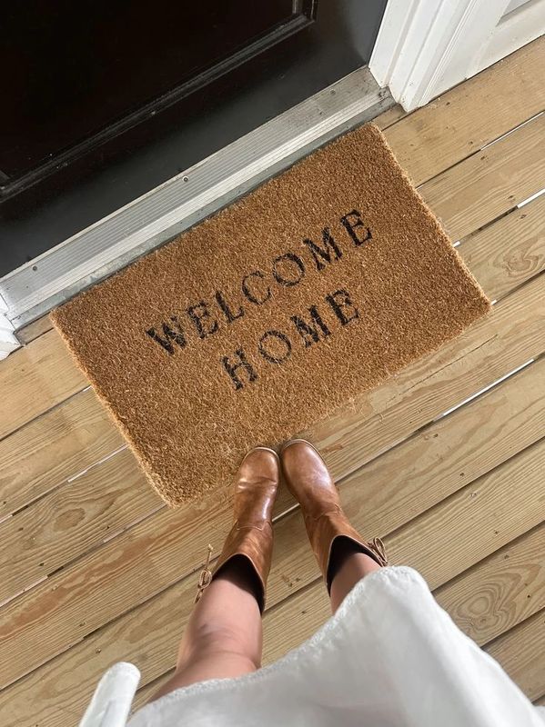 Person wearing brown boots standing on a wooden porch with a 'Welcome Home' doormat.