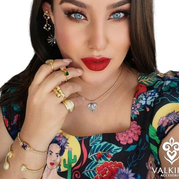 Woman with blue eyes, red lipstick, and colorful floral dress adorned with gold jewelry.