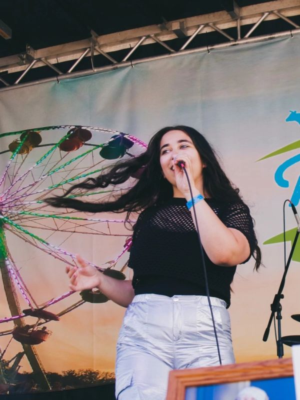 Woman singing on stage with a Ferris wheel backdrop at sunset.