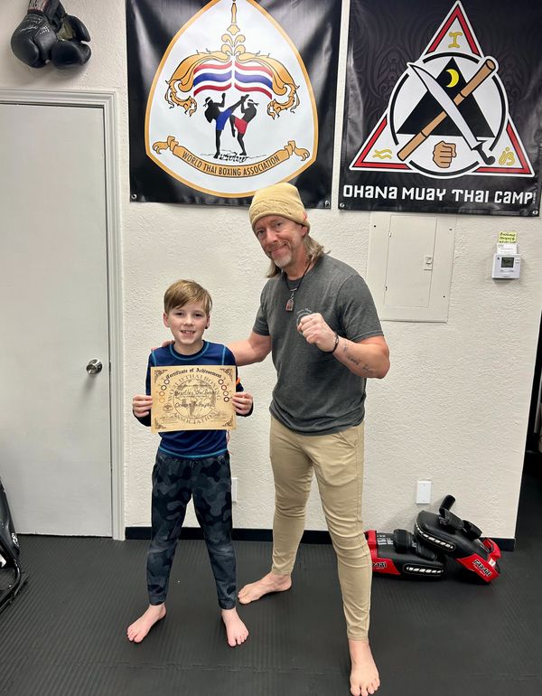 Boy proudly holds a certificate with his Muay Thai instructor in a gym.