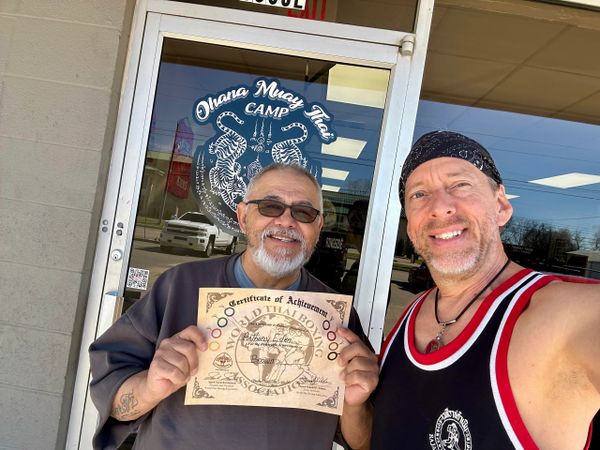 Two men smiling outside Ohana Muay Thai Camp, one holding a certificate.