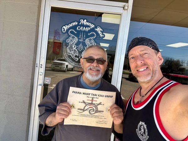 Two men smiling, one holding a Muay Thai promotion certificate outside a martial arts gym.