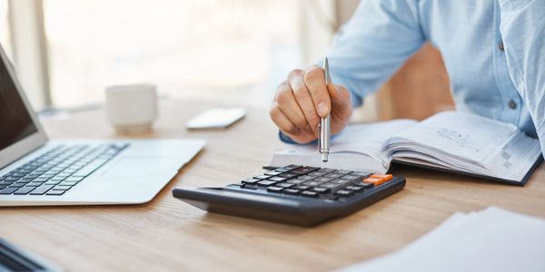 Person using a calculator and pen with a laptop and notebook on desk.