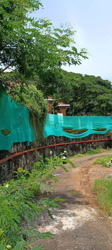 Winding dirt path alongside green mesh fence and lush trees.