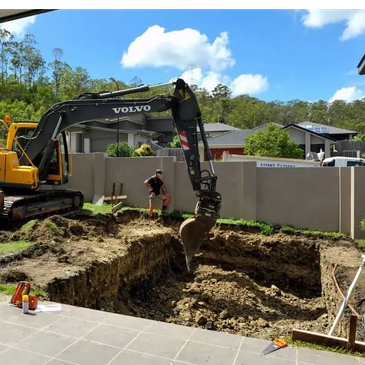Excavator digging a large hole in a backyard with a person standing nearby.