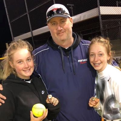 Coach with two smiling girls holding softball medals and a ball at night field.