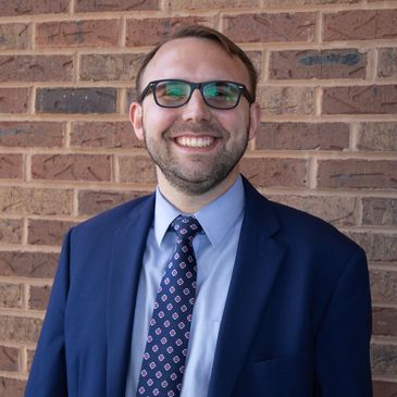 Smiling man in a blue suit and patterned tie stands against a brick wall.