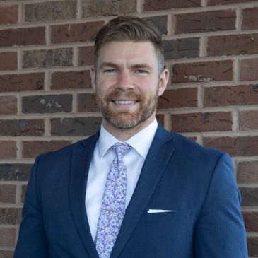 Man in blue suit and floral tie smiling against a brick wall.