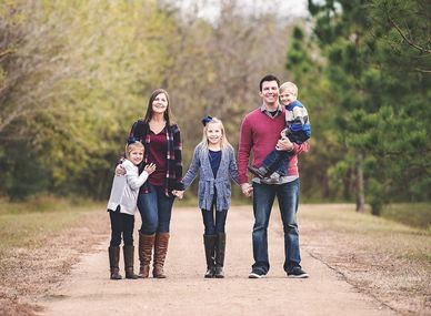 A happy family of five posing outdoors on a dirt path surrounded by trees.