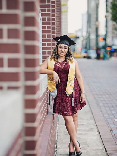 A woman in a graduation cap and sash poses against a brick wall.