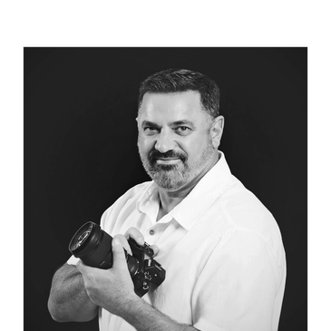 Portrait of a man holding a Nikon camera, dressed in a white shirt, against a black background.