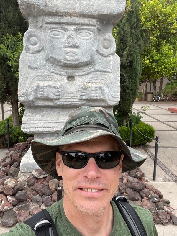 Man wearing sunglasses and a camouflage hat poses in front of an ancient stone statue.