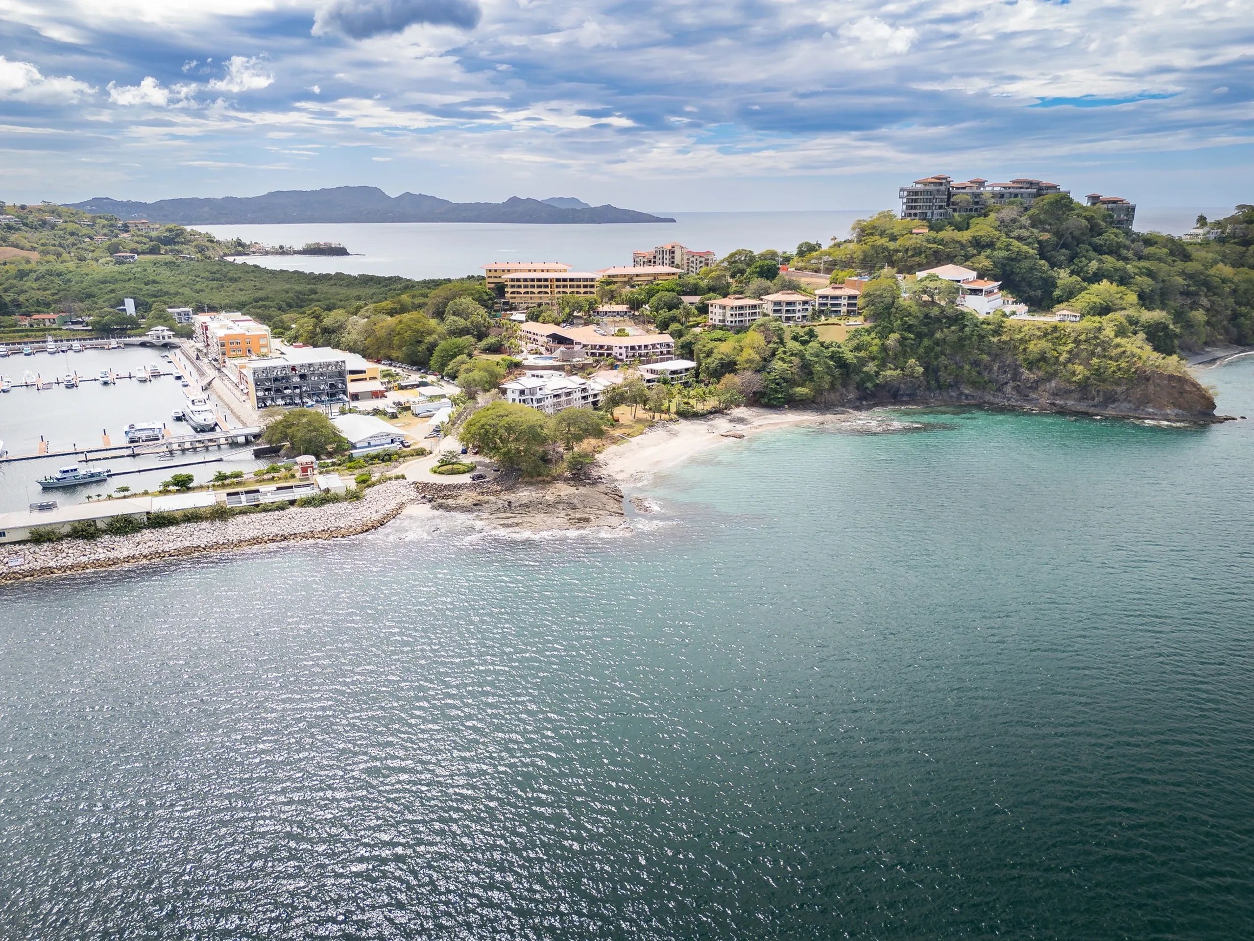 Coastal town with buildings, greenery, marina, and mountains under a cloudy sky.
