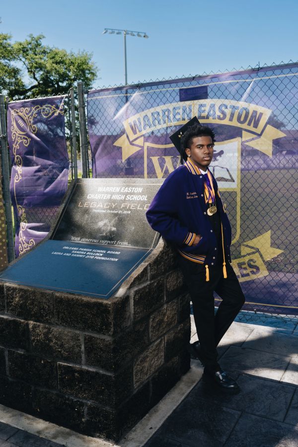 Graduate in cap and gown posing by Warren Easton Charter High School plaque.