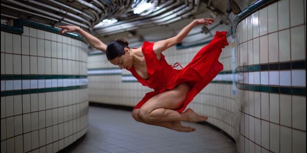 Dancer in a red dress leaps gracefully in a tiled corridor.