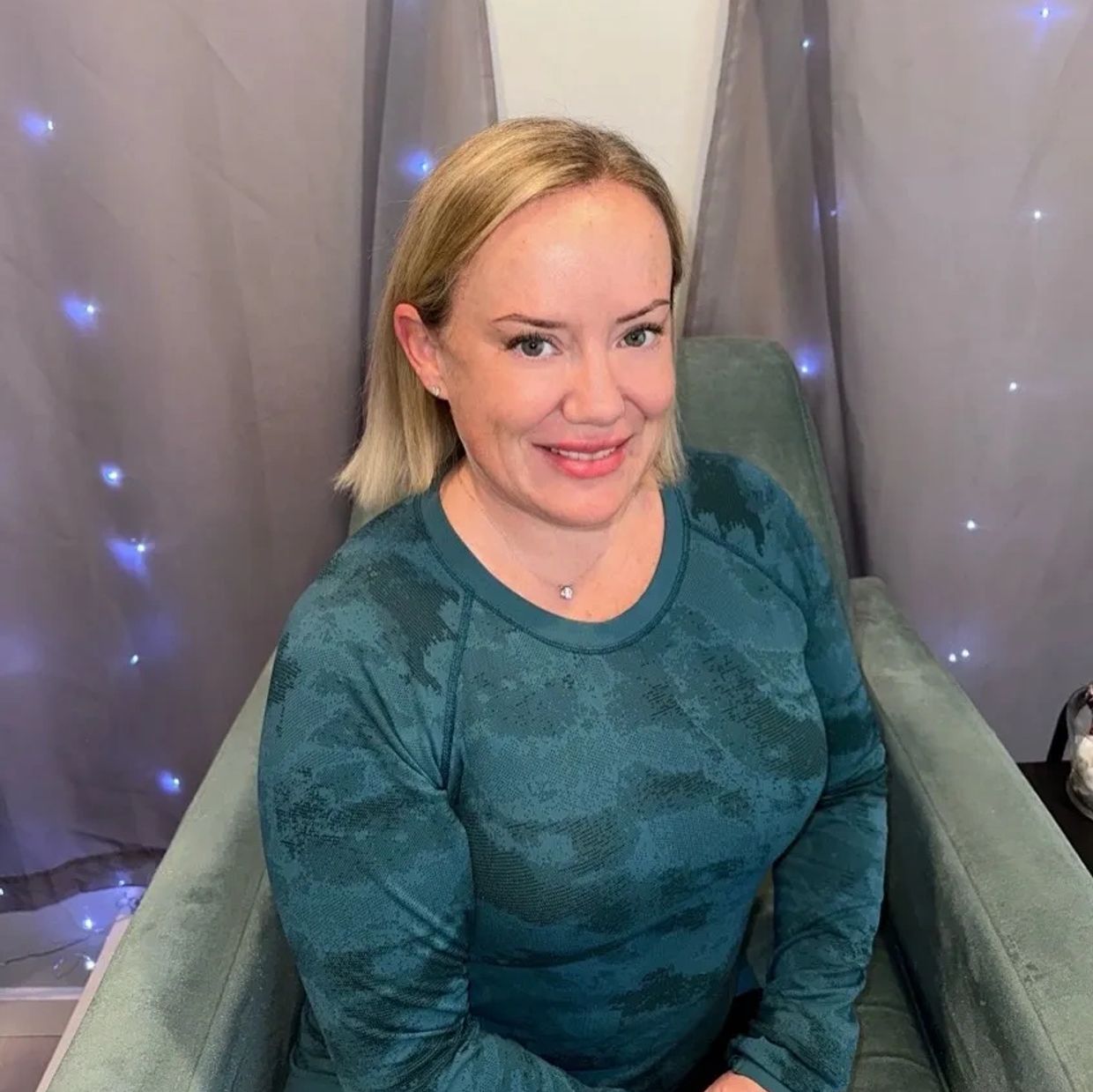Smiling woman in a teal shirt sitting on a chair with fairy lights in the background.