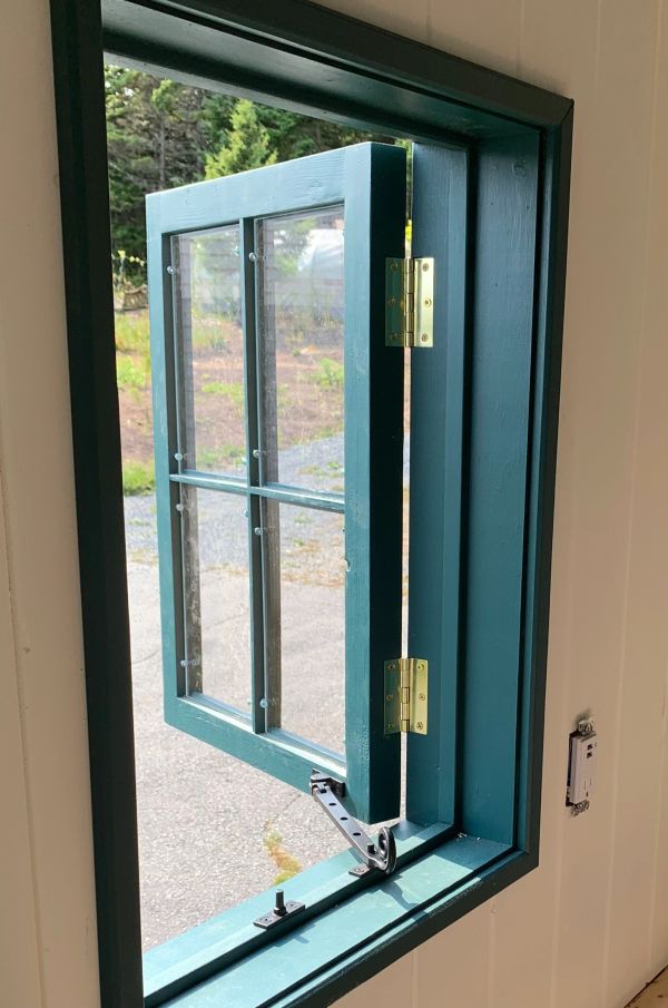 Teal-painted wooden casement window with brass hinges open to a sunny outdoor scene.