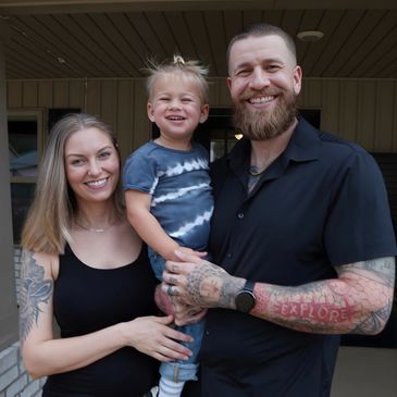A smiling family of three posing outdoors, showcasing tattoos and joyful expressions.