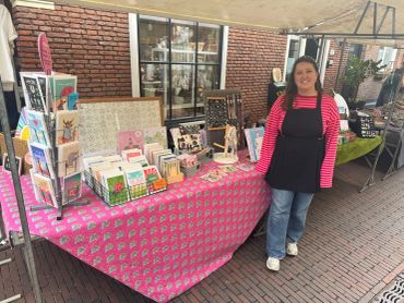 Vrouw poseert bij marktkraam met kaarten en sieraden.
