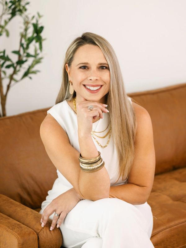 Smiling woman in white outfit sitting on a brown couch with gold accessories.