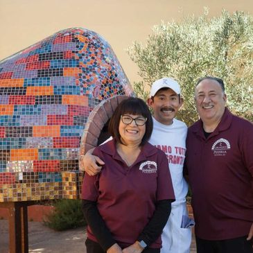Three people smiling in front of a colorful mosaic oven.