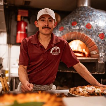 Man in a maroon shirt and cap presenting freshly baked pizzas.
