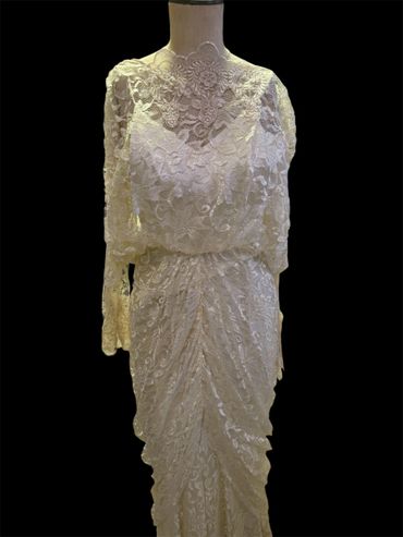 Elegant white lace dress with long sleeves on a mannequin against a black background.