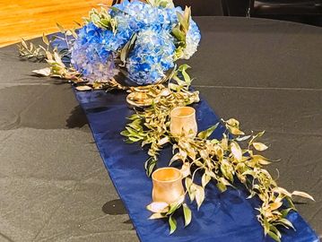 Centerpiece featuring blue hydrangea and gold accents on a navy table runner and black tablecloth.