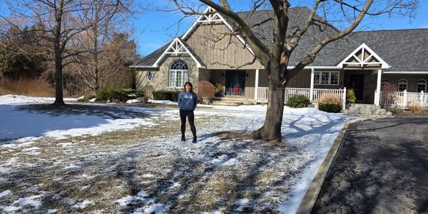 Woman standing in front of a house with snow on the ground and bare trees.
