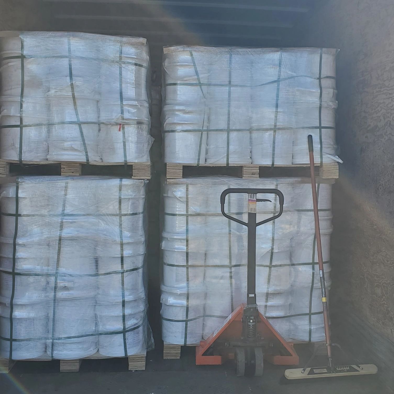 Four large pallets wrapped in plastic inside a truck with a pallet jack and broom.