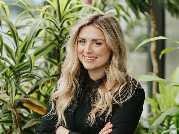 Smiling woman with long blonde hair standing in front of green plants.