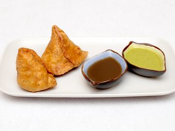 Two samosas with green and tamarind chutneys on a white plate.