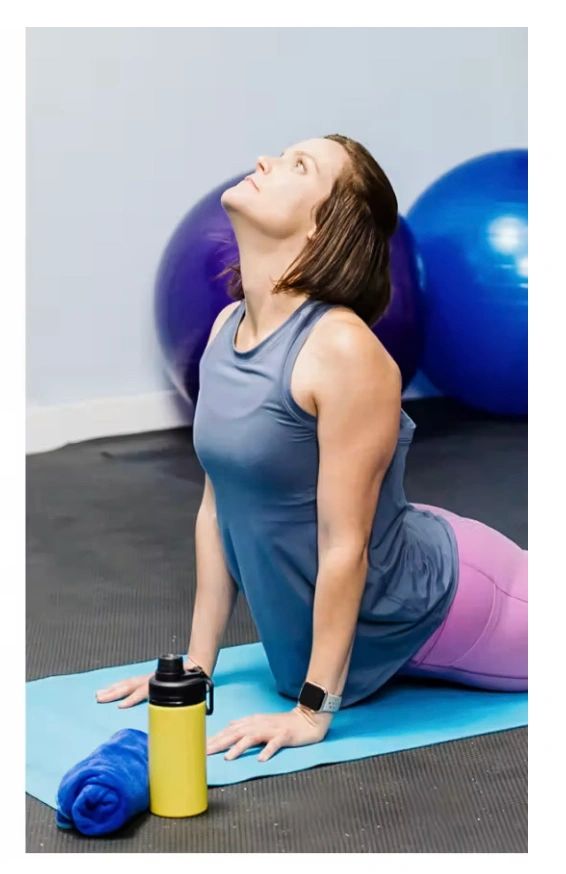 Woman stretching on yoga mat in gym with water bottle and towel nearby.