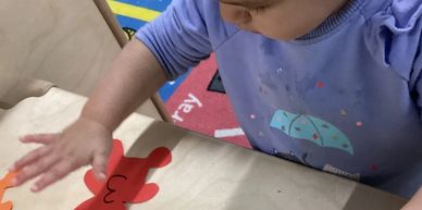 A child in a purple shirt playing with a red bear cutout on a table.