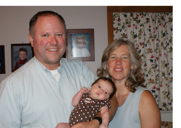 Man and women standing holding infant with pictures two other children in the background