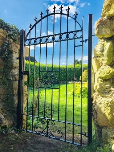 Ornate black iron gate in a stone wall opening to a sunny garden.