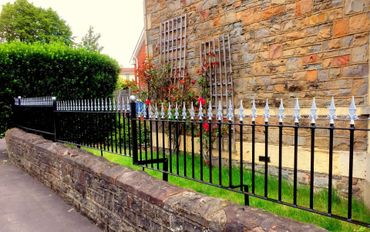 Black metal fence with pointed tops along a stone wall and garden.