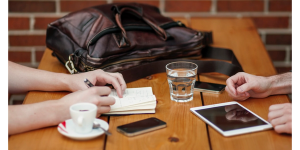 arms of people at a wooden table with bag, espresso, water, iPad, iPhone, notebook & pen to hand