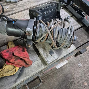 A winch with coiled steel cable on a wooden surface next to gloves.