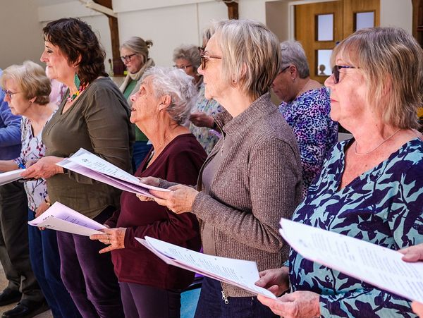 A group of elderly women singing together, holding sheet music.