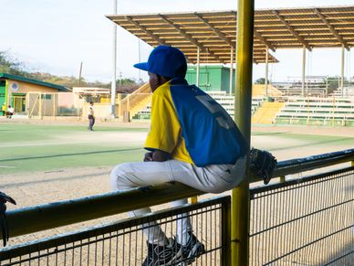A young baseball player sits on a fence watching the game at an empty field.