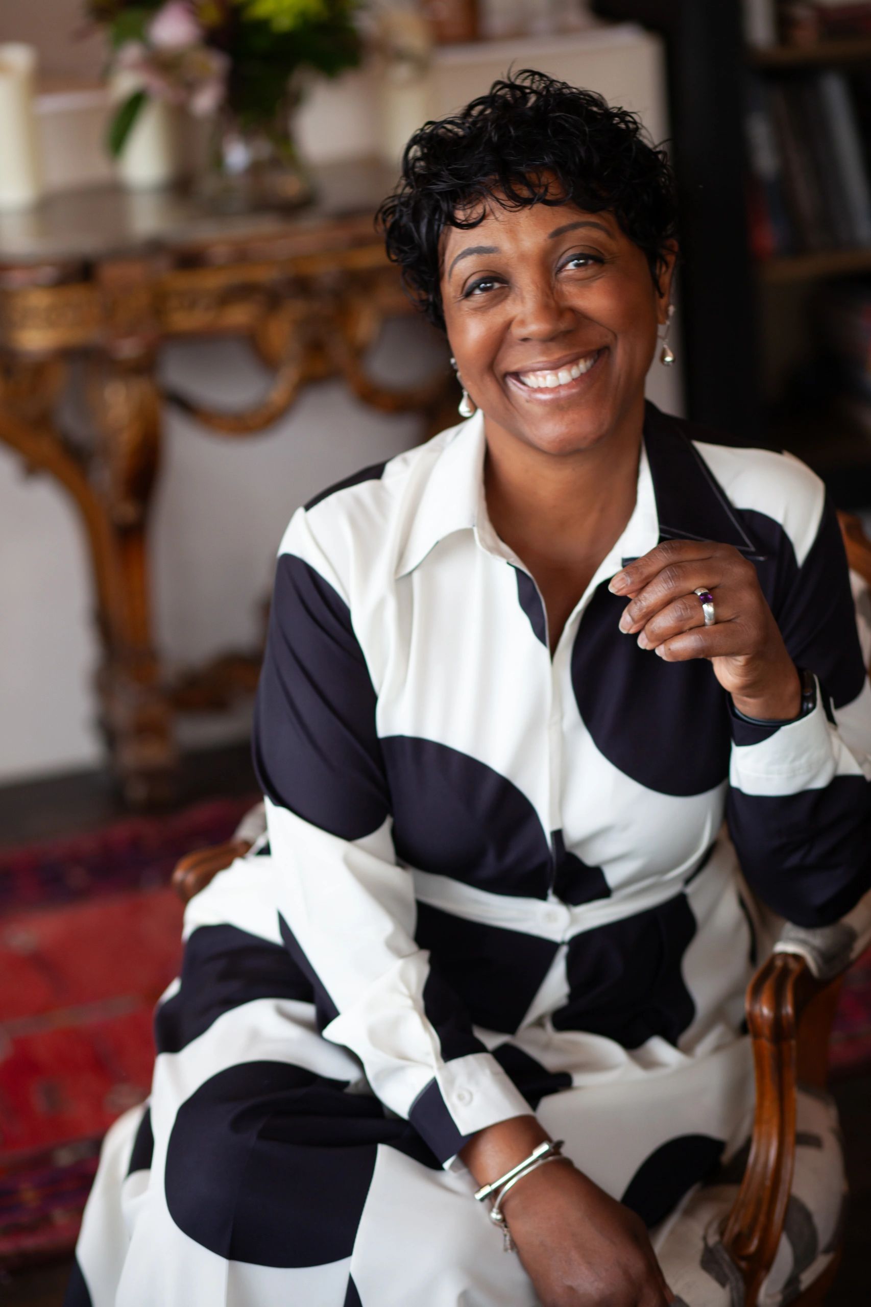 Smiling woman in a black and white dress sitting on a chair in a cozy room.