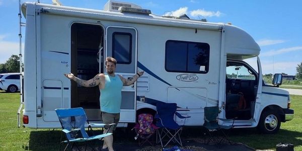 Man standing in front of a white RV with camping chairs on grass.