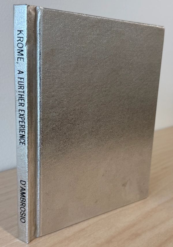 Silver-covered book titled 'Krome A Further Experience' by D'Ambrosio on a wooden surface.