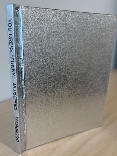 Silver metallic book titled "You Dress 'Funny' An Experience" by D'Ambrosio.