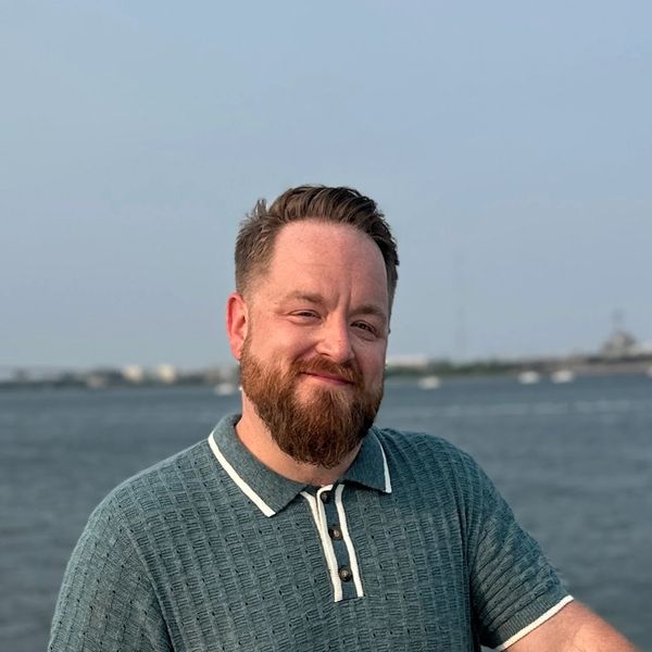 Smiling man with a beard standing by a waterfront on a clear day.